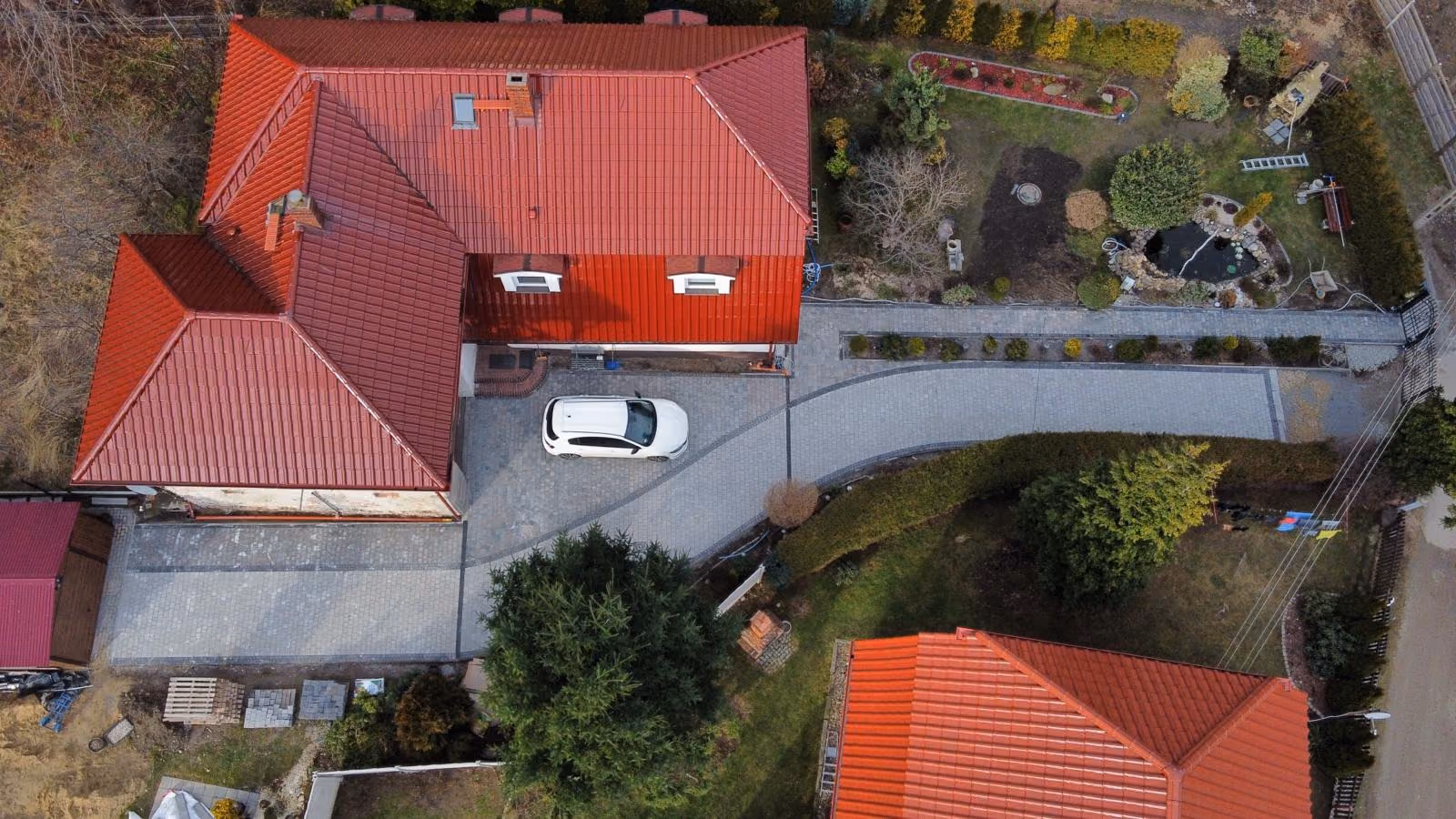Widok z drona: podjazd z szarej kostki od bramy wzdłuż żywopłotu do garażu i placu manewrowego przed domem