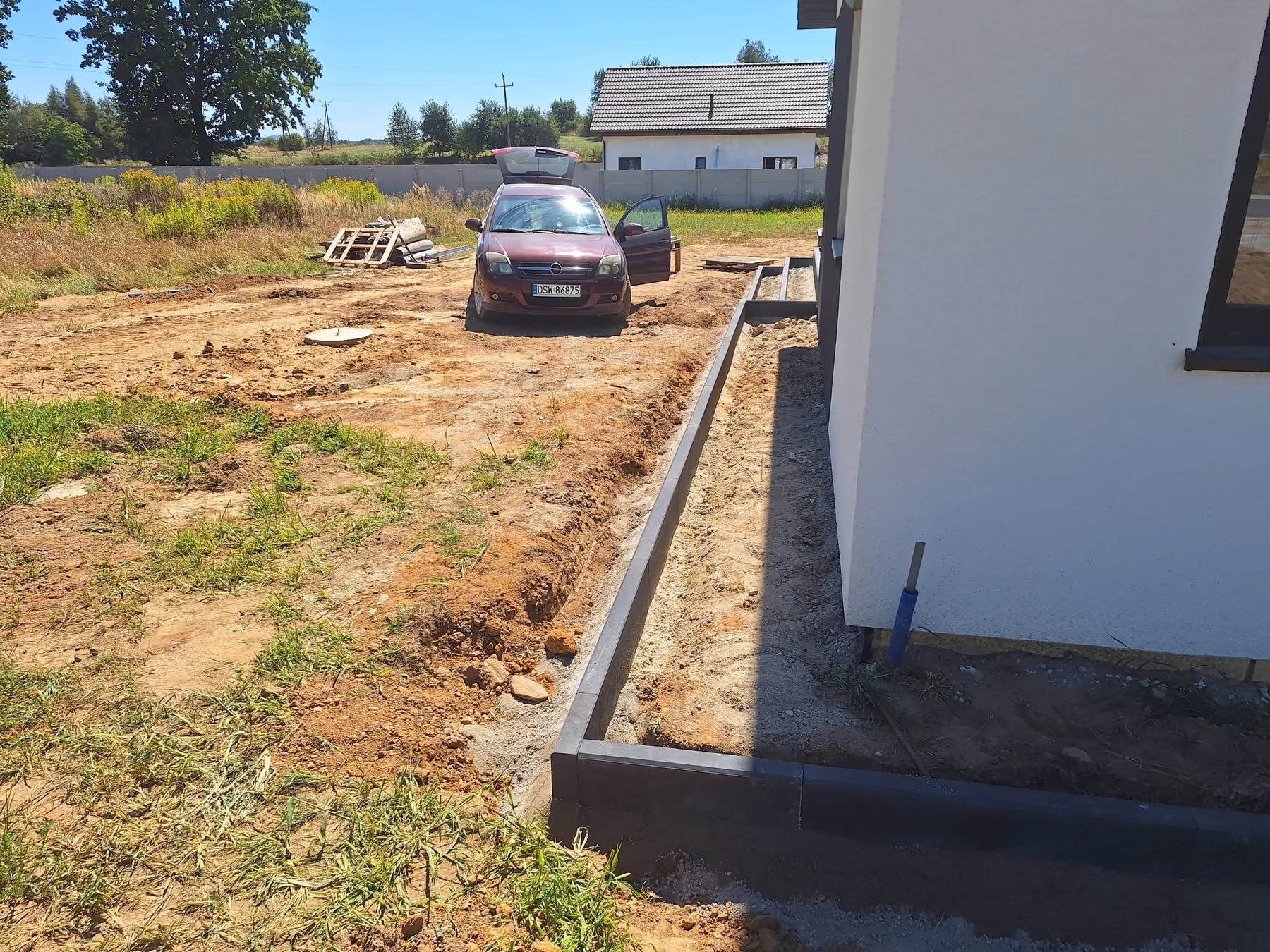 Ciemne obrzeże na krawędzi nawierzchni przy narożniku domu, obok wykop pod opaskę
